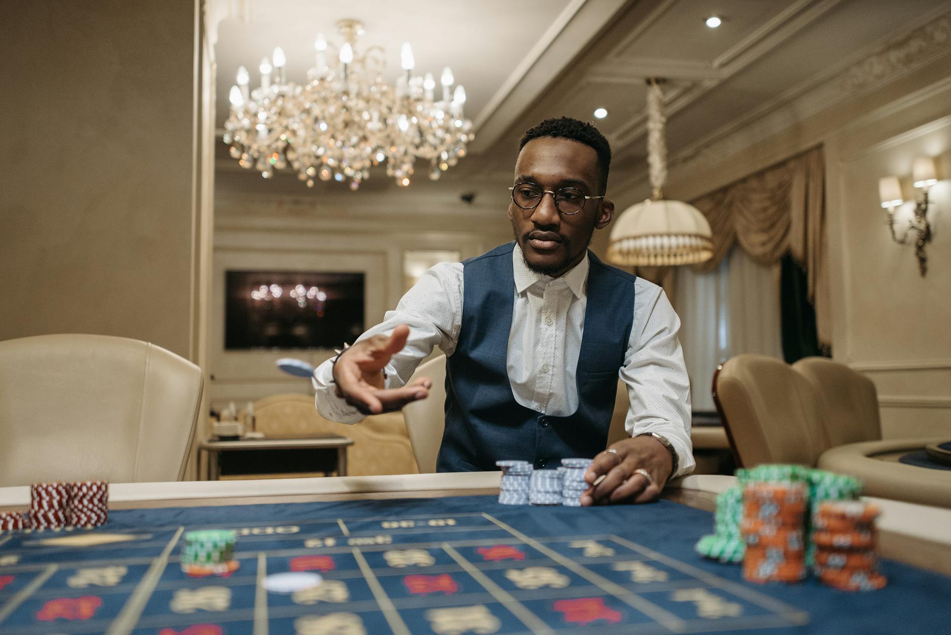 A man in formal attire plays roulette in a luxurious casino under a chandelier.