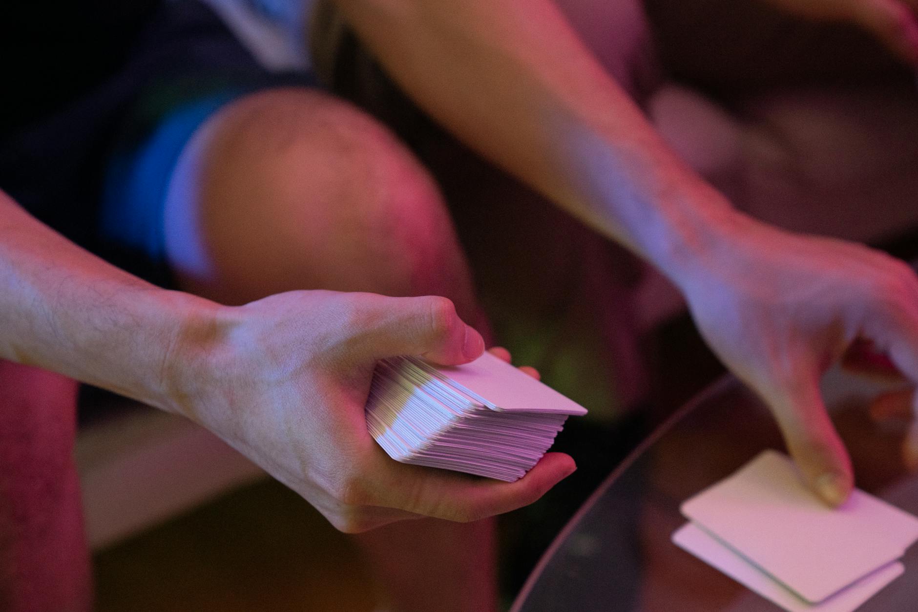 Close-up of hands shuffling a deck of cards on a glass table with pink lighting.